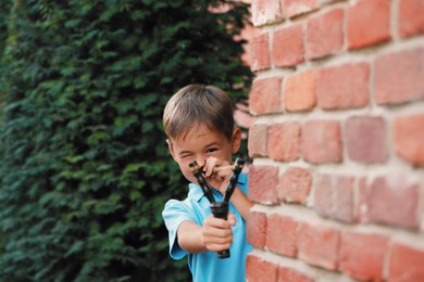Little boy playing with slingshot near brick wall outdoors