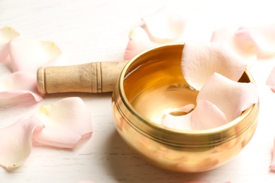 Photo of Golden singing bowl with petals and mallet on white wooden table, closeup. Sound healing