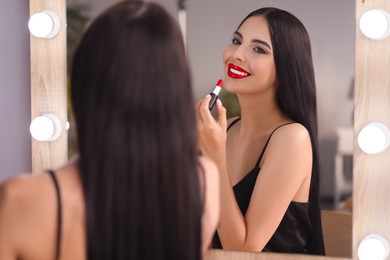 Photo of Young woman applying beautiful red lipstick in front of mirror indoors