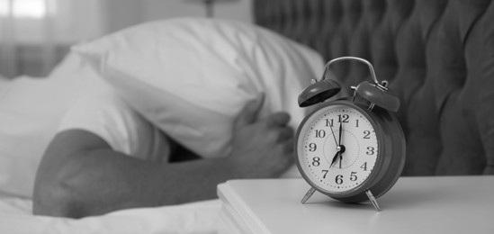 Image of Alarm clock and man covering head with pillow in bedroom, selective focus. Black and white photography