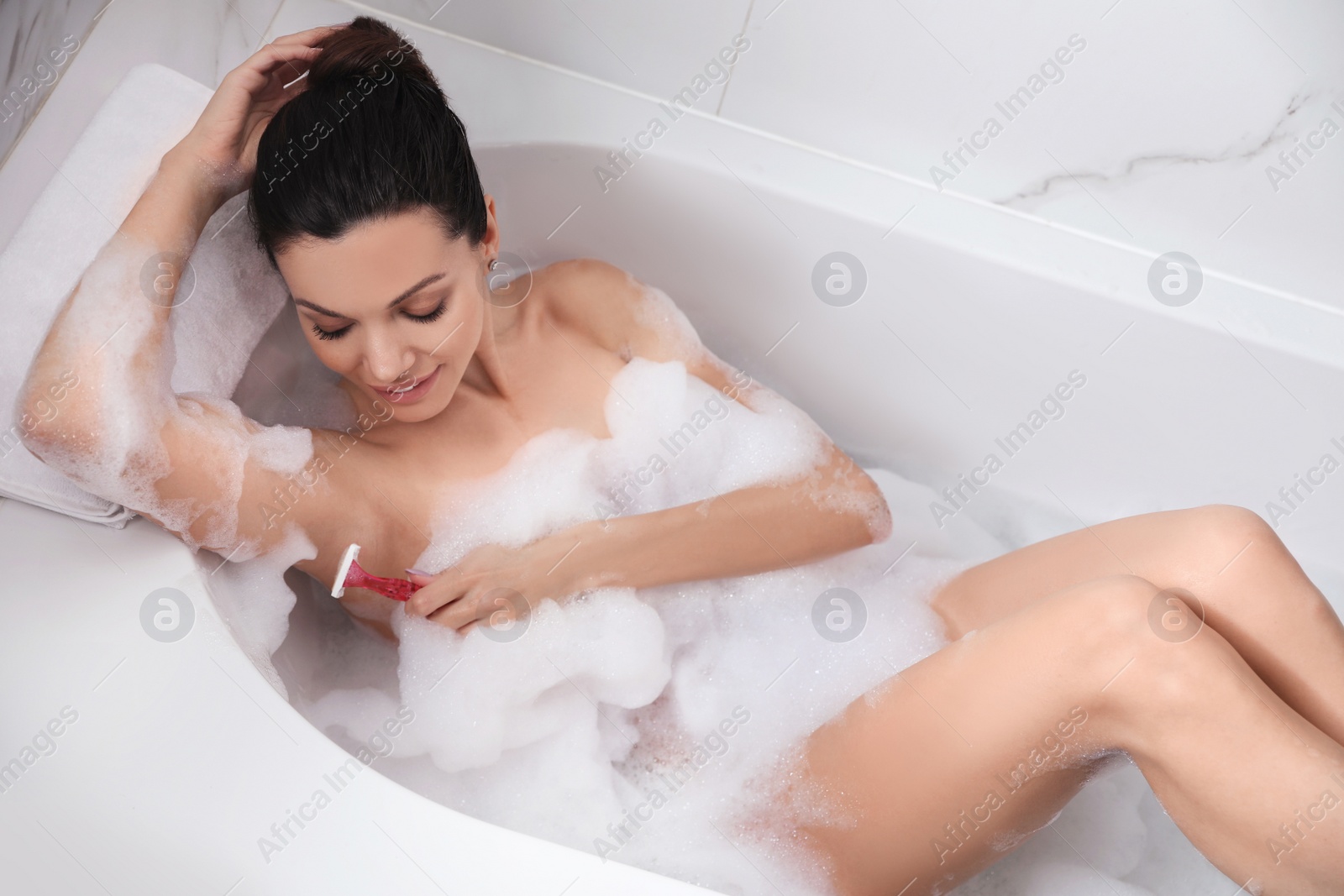 Photo of Young woman shaving armpit while having bubble bath, above view