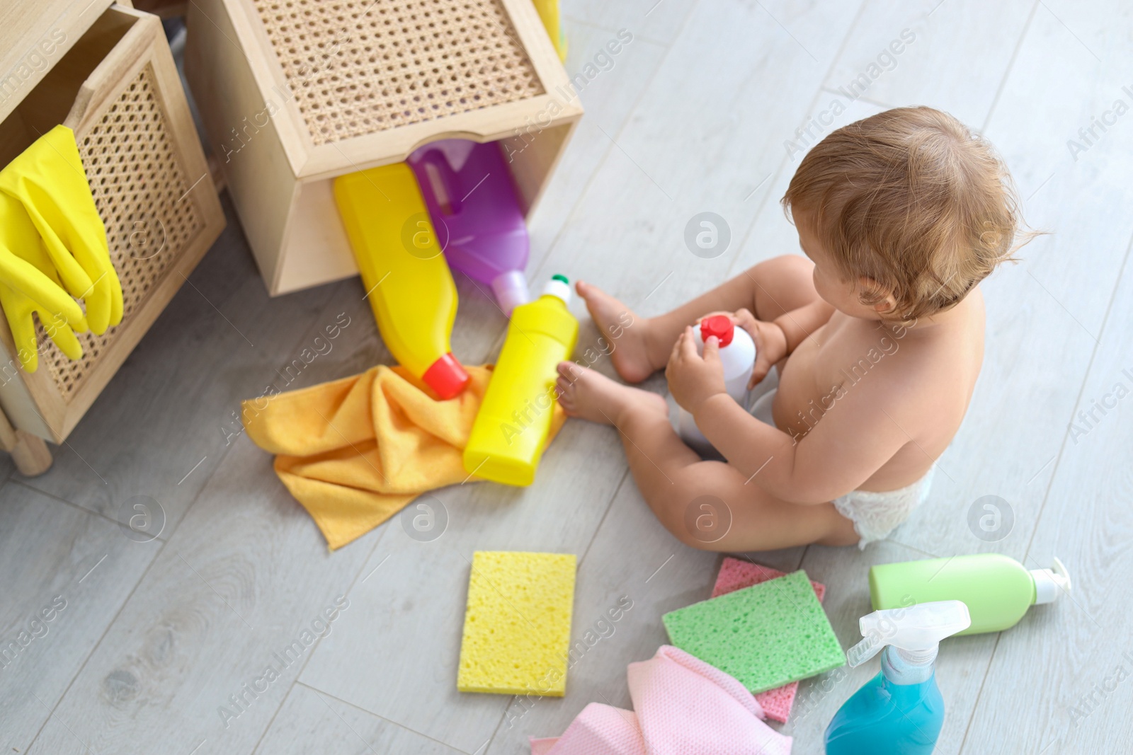 Photo of Cute baby playing with bottle of detergent on floor at home, above view. Dangerous situation