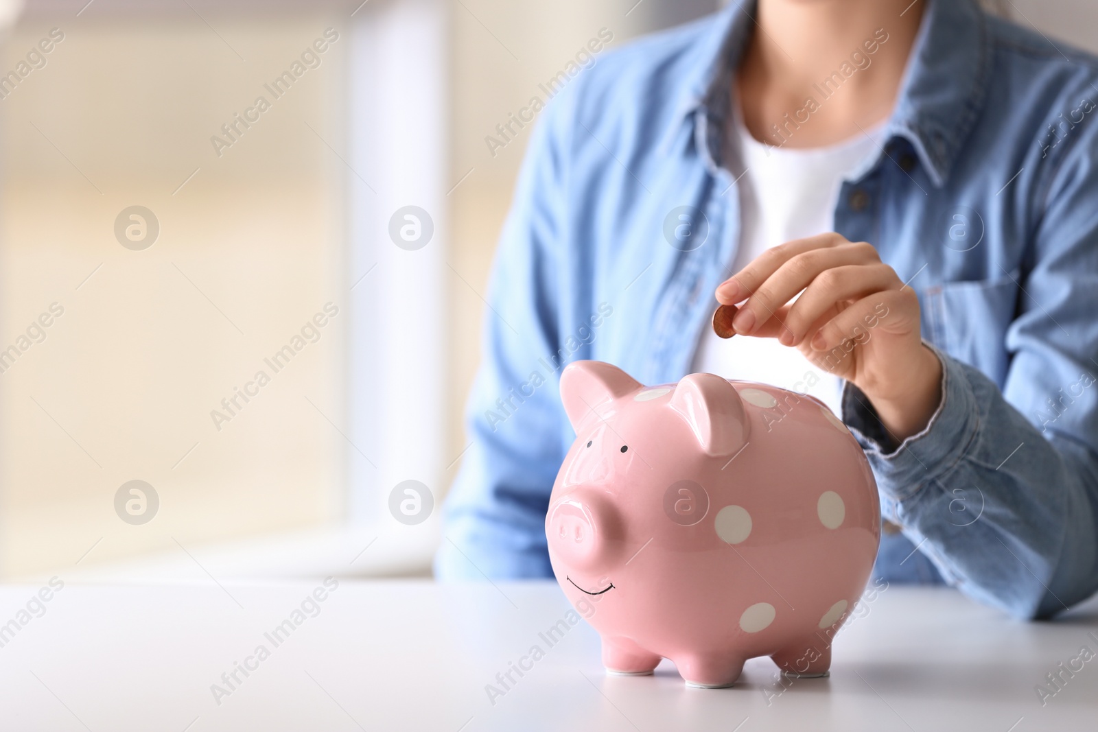 Photo of Woman putting coin into piggy bank at table indoors, closeup. Space for text