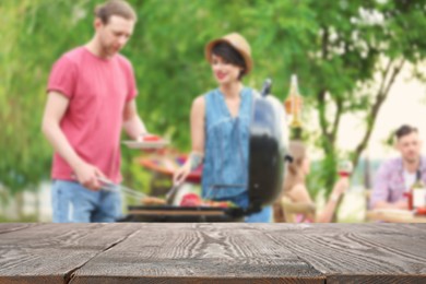 Image of Empty wooden table and blurred view of people having barbecue with modern grill outdoors