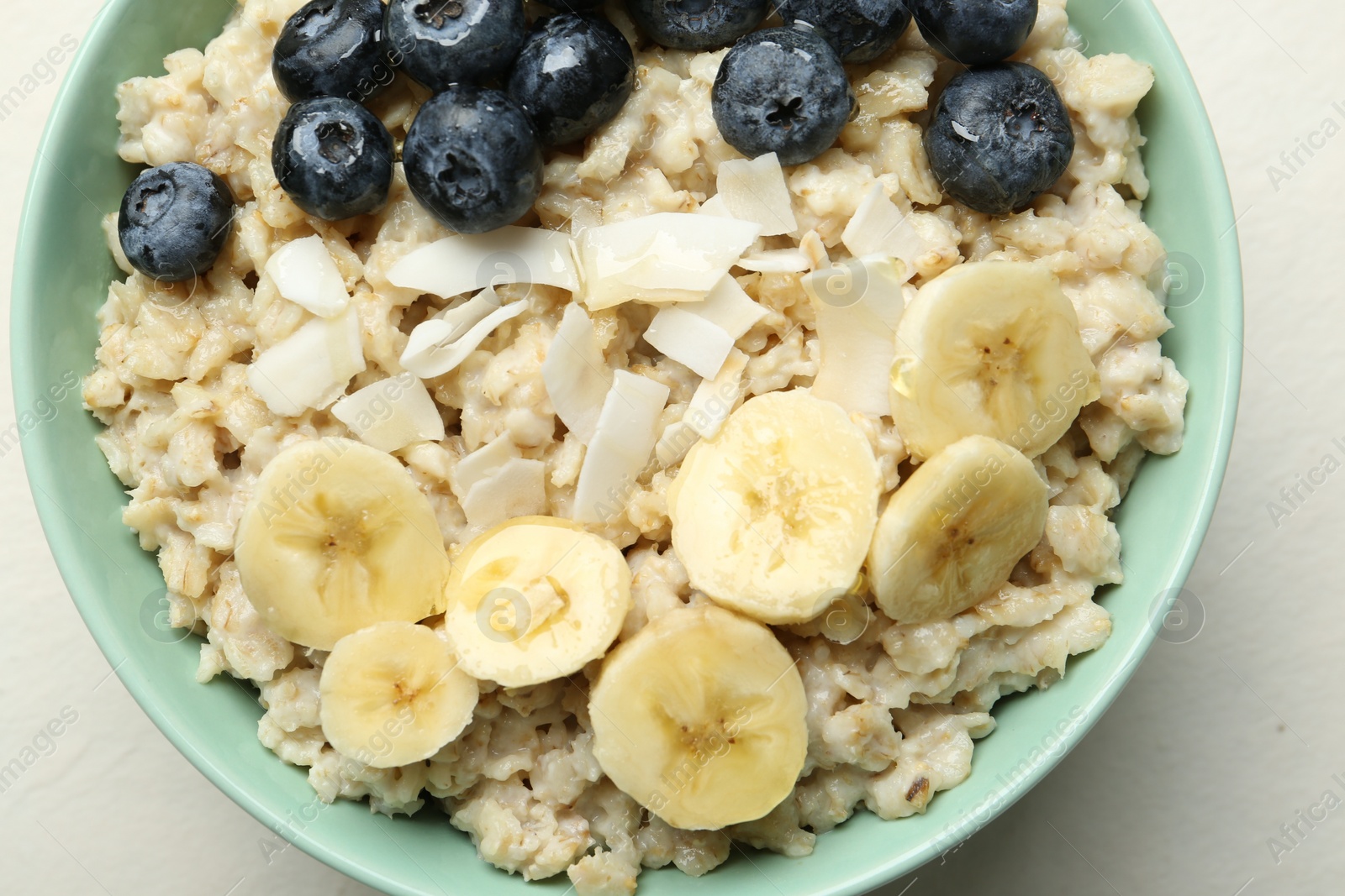 Photo of Tasty oatmeal with banana, blueberries, coconut flakes and honey served in bowl on beige table, top view