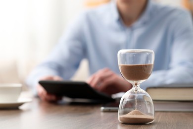 Photo of Hourglass with flowing sand on desk. Man using calculator indoors, selective focus