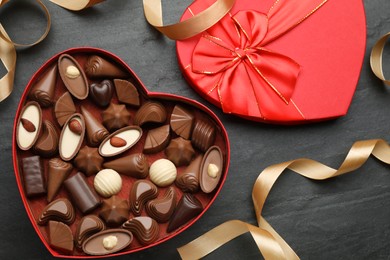 Photo of Heart shaped box with delicious chocolate candies and ribbon on black table, flat lay