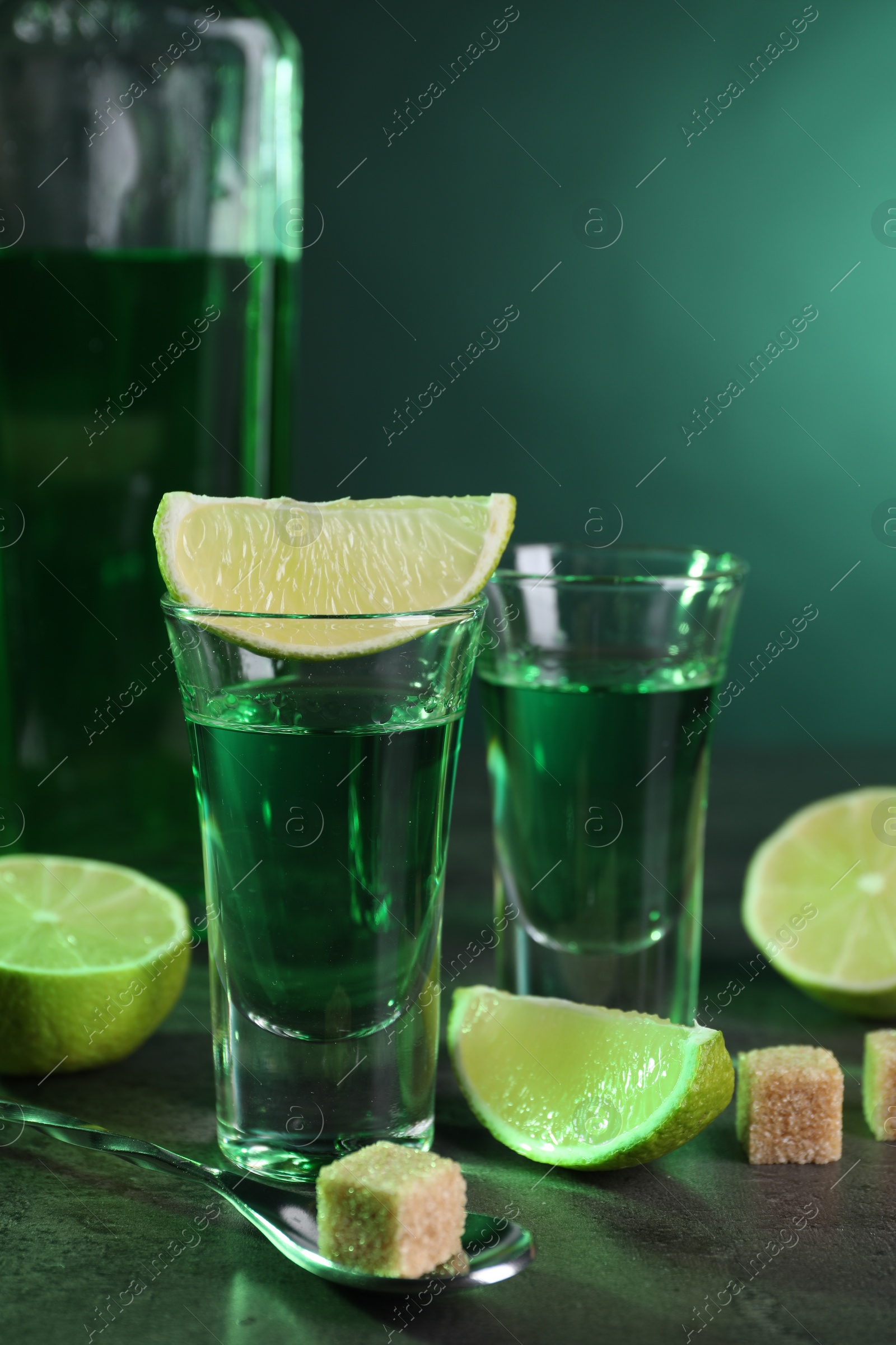 Photo of Absinthe in shot glasses, spoon, brown sugar cubes and lime on gray textured table, closeup. Alcoholic drink