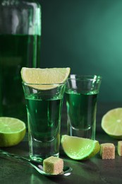 Absinthe in shot glasses, spoon, brown sugar cubes and lime on gray textured table, closeup. Alcoholic drink