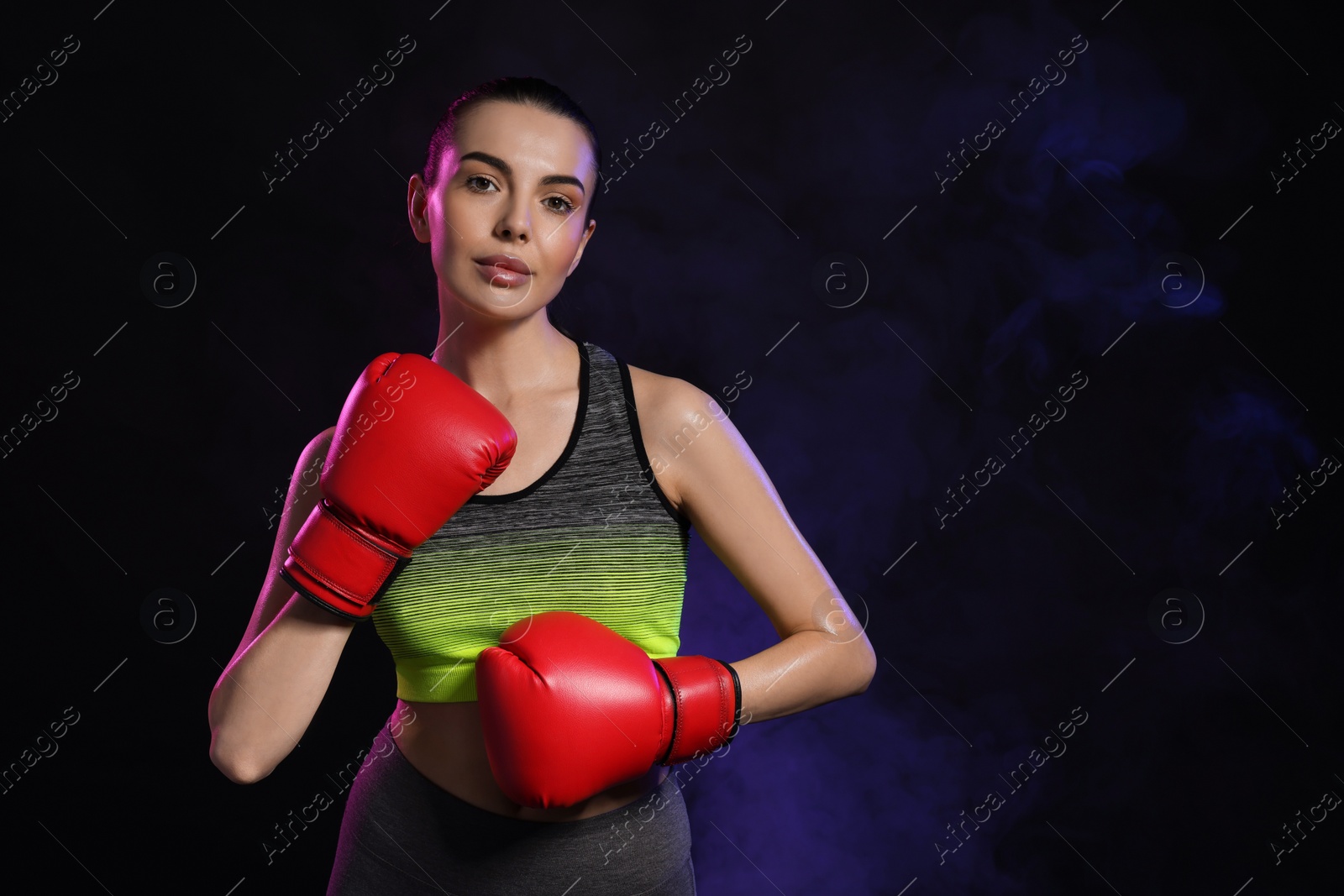 Photo of Portrait of beautiful woman wearing boxing gloves in color lights on black background. Space for text