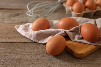 Photo of Raw chicken eggs, napkin, board and whisk on wooden table, closeup