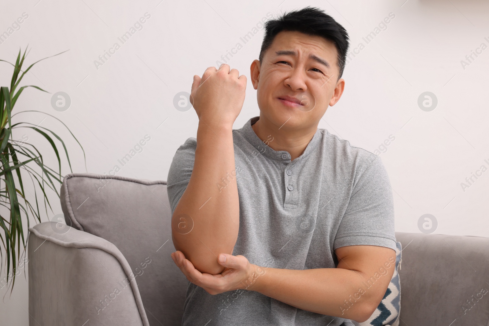 Photo of Asian man suffering from pain in his elbow on armchair indoors