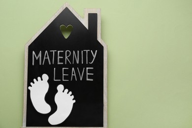 Photo of Wooden house figure with words Maternity Leave and paper cutout of baby feet on pale green background, top view. Space for text