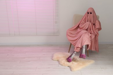 Glamorous ghost. Woman in pink sheet and high heel shoes on armchair indoors, space for text