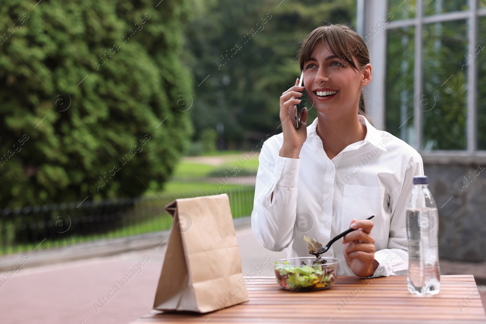 Photo of Happy businesswoman talking on smartphone while having lunch at wooden table outdoors, space for text