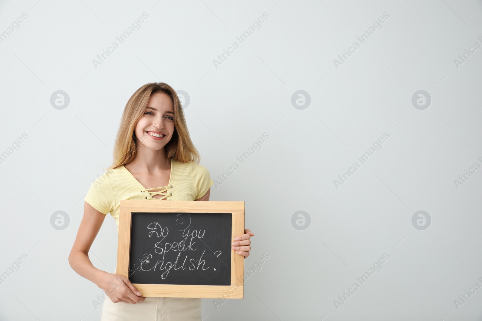 Photo of Young female teacher holding chalkboard with words DO YOU SPEAK ENGLISH? on light background. Space for text