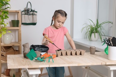 Little girl adding soil into peat pots at wooden table in room. Growing vegetable seeds