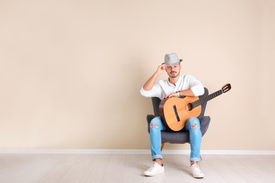 Photo of Handsome young man with guitar in armchair near color wall indoors. Space for text