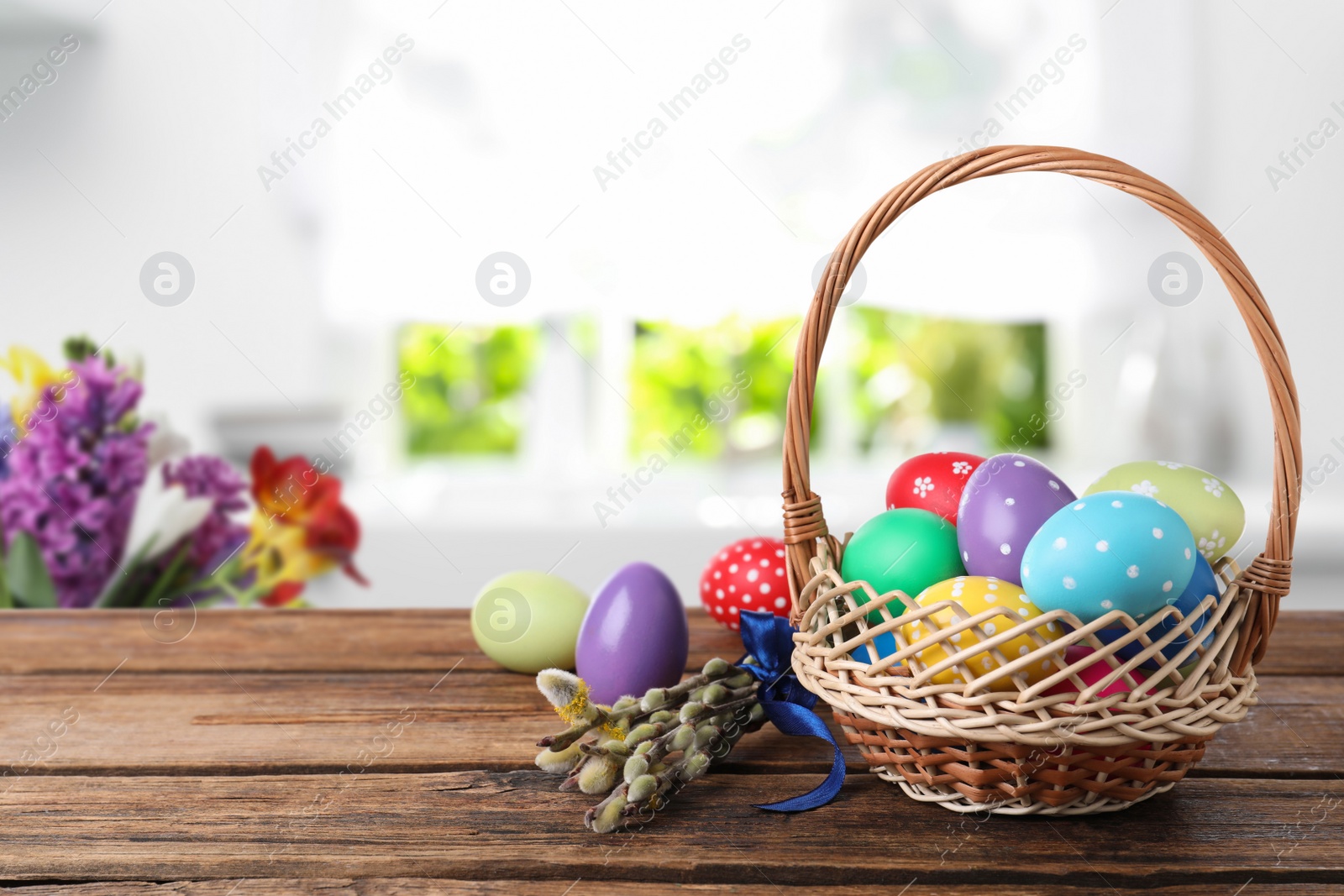 Image of Colorful Easter eggs in wicker basket and willow branches on wooden table indoors, space for text