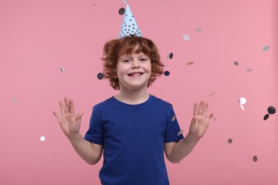 Photo of Happy little boy in party hat under falling confetti on pink background