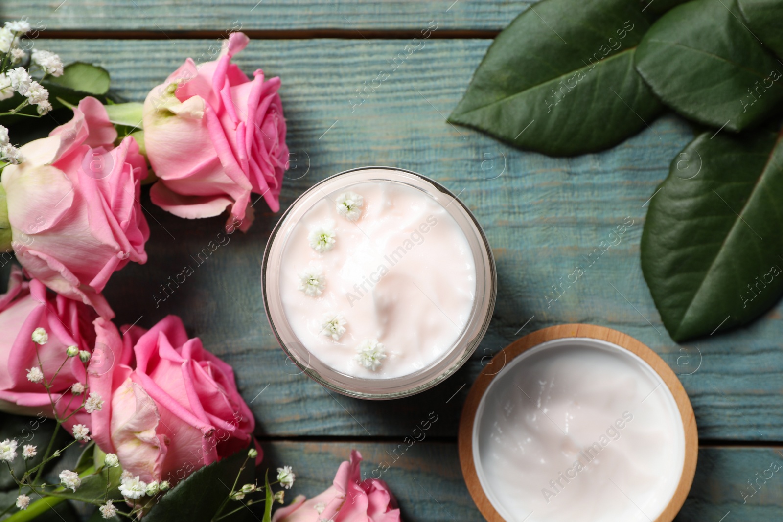 Photo of Jar of face cream and pink roses on light blue wooden table, flat lay