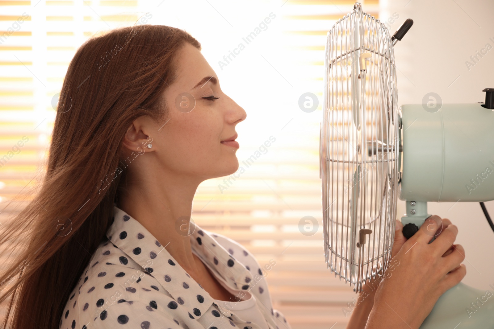 Photo of Woman enjoying air flow from fan near window indoors. Summer heat