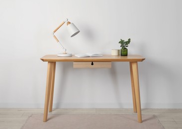 Photo of Stylish modern desk lamp, open book and plant on wooden table near white wall indoors