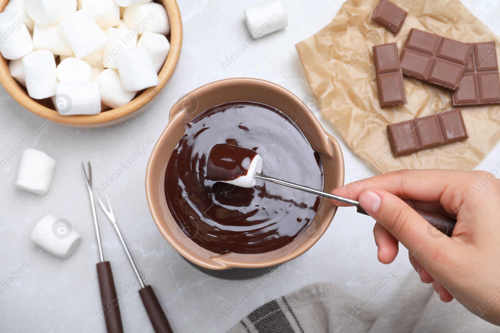 Photo of Woman dipping marshmallow into melted chocolate at light grey marble table, above view