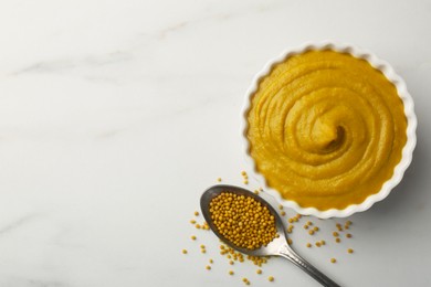 Photo of Tasty mustard sauce in bowl and spoon with dry seeds on white marble table, flat lay. Space for text