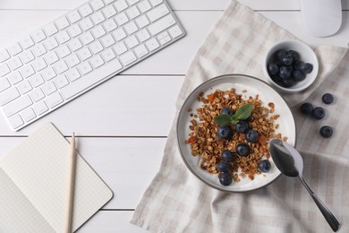 Photo of Delicious granola with blueberries in bowl, stationery and computer keyboard on white wooden table, flat lay