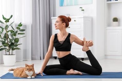 Photo of Beautiful woman with cute red cat practicing yoga on mat at home