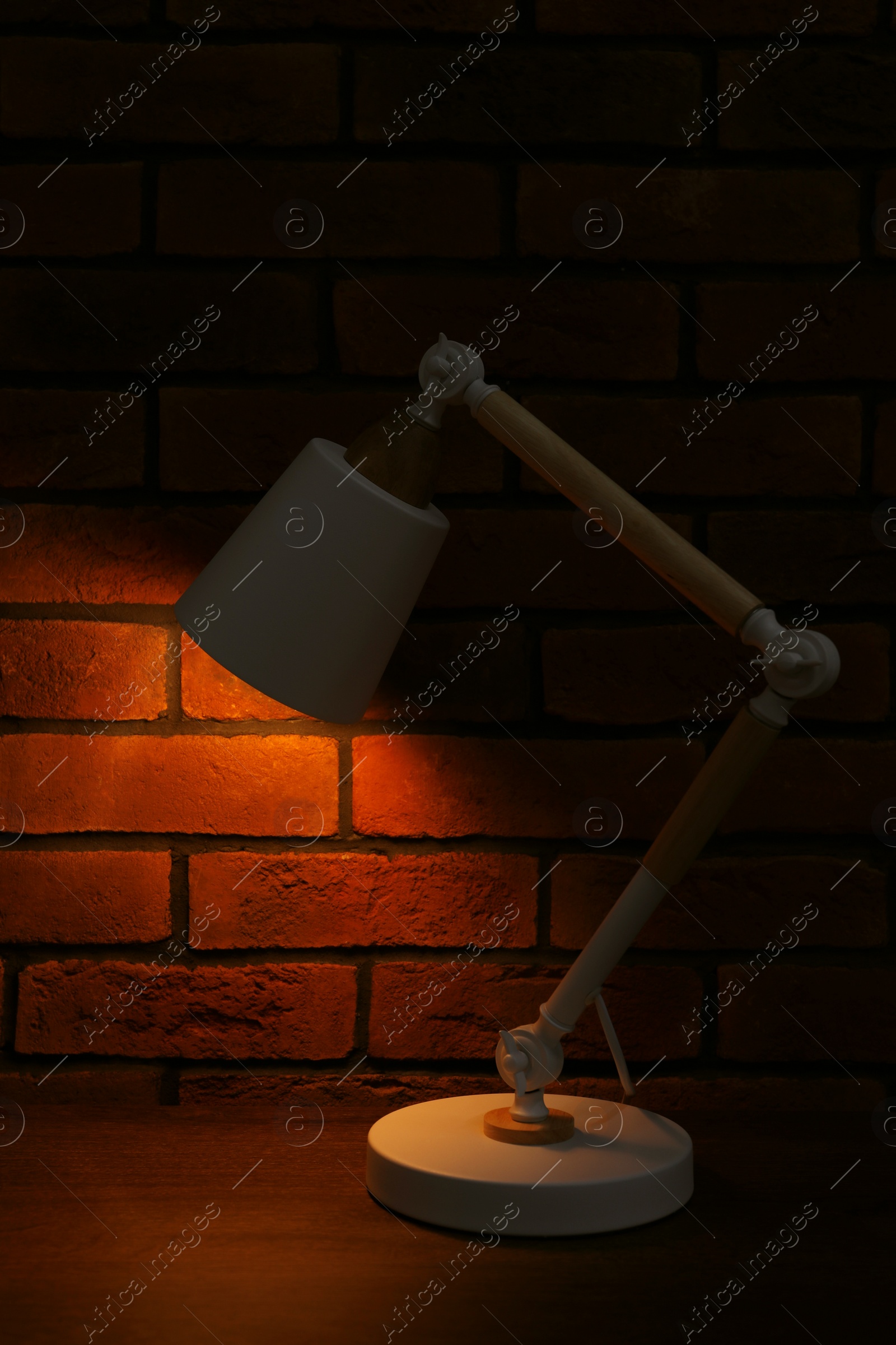 Photo of Stylish modern desk lamp on wooden table near brick wall at night