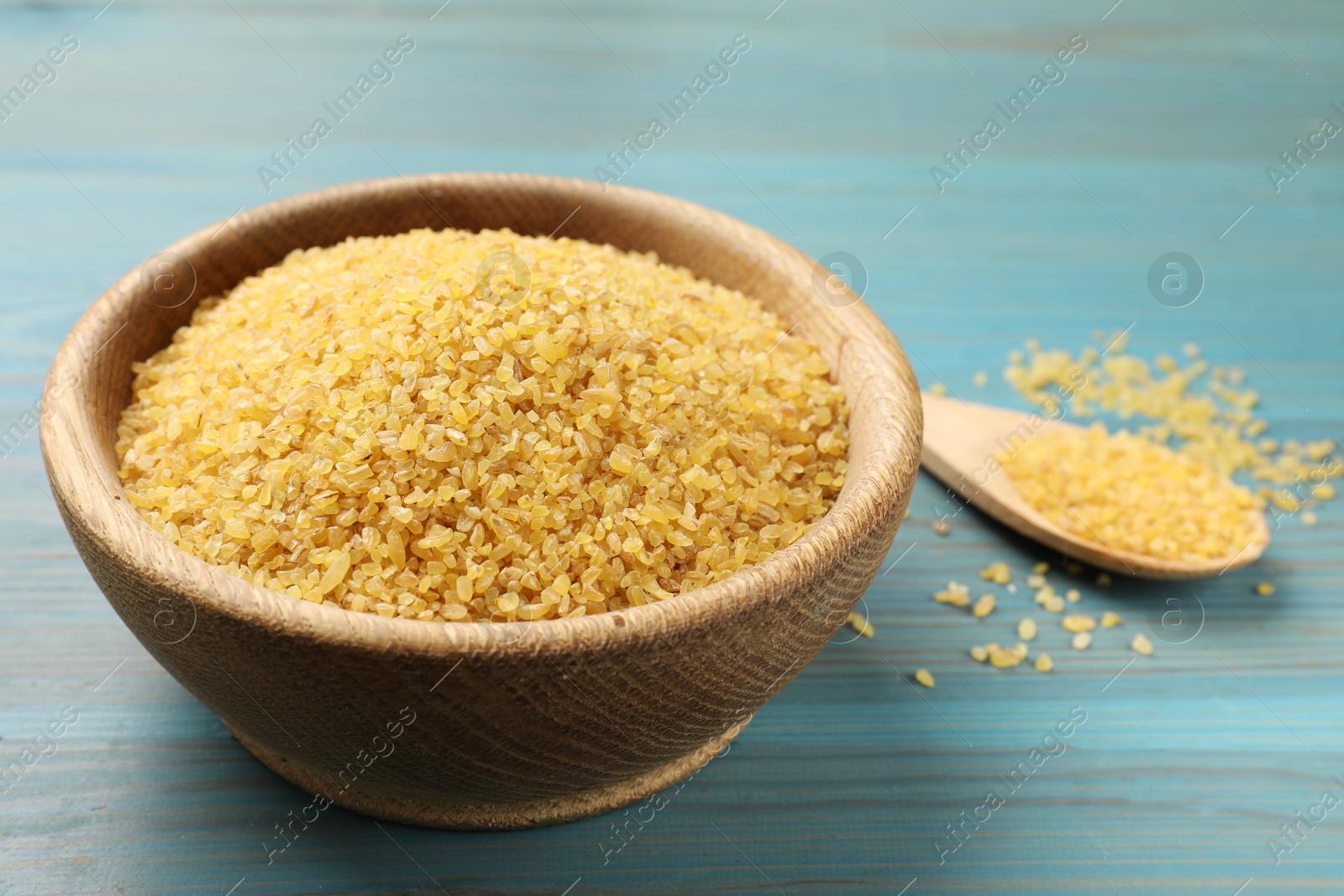 Photo of Bowl and spoon with raw bulgur on light blue wooden table, closeup