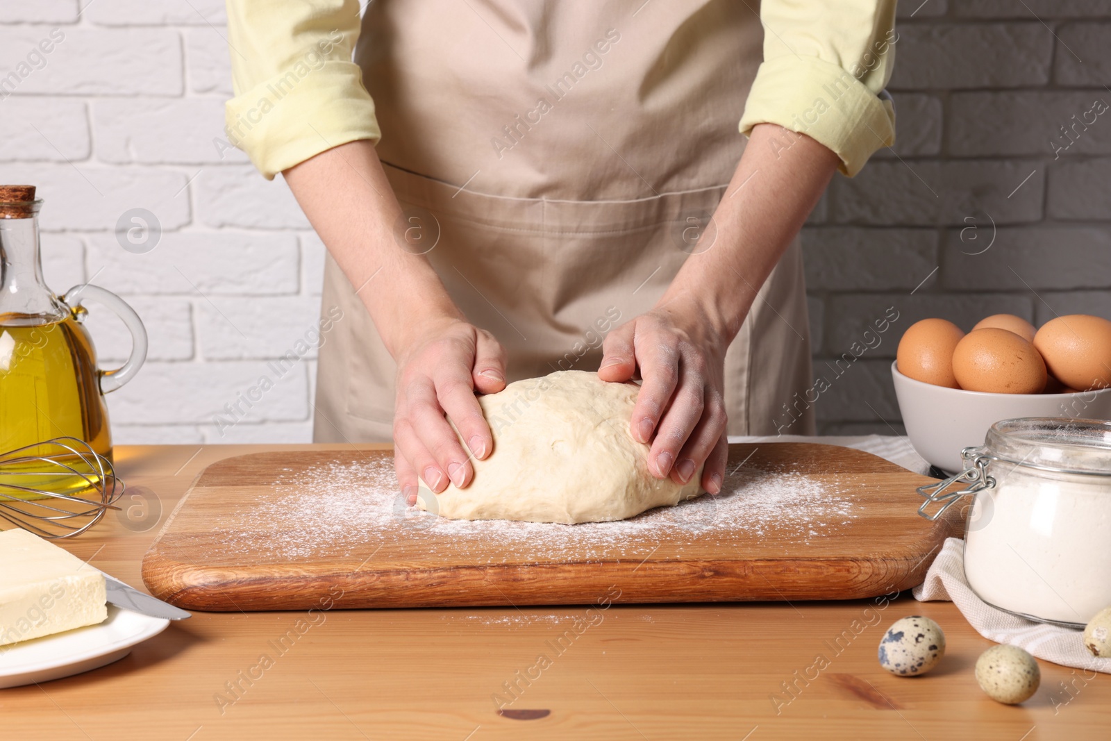 Photo of Woman kneading dough at wooden table near white brick wall, closeup