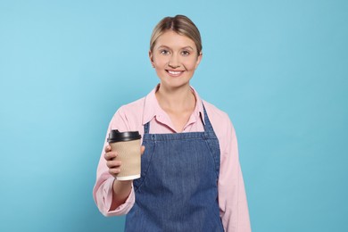 Photo of Beautiful young woman in denim apron with cup of coffee on light blue background