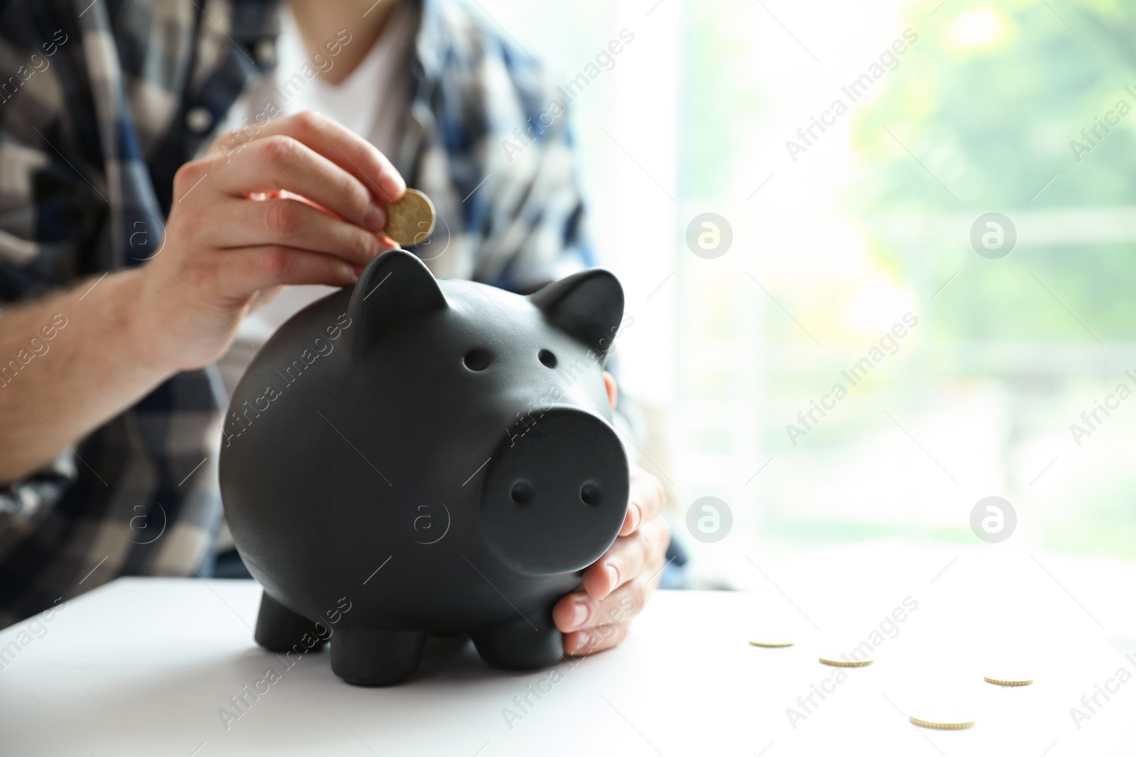Photo of Man putting coin into piggy bank at white table against blurred background, closeup