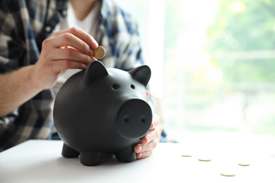 Man putting coin into piggy bank at white table against blurred background, closeup