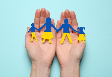 Image of Woman holding paper family figures in colors of Ukrainian flag on light blue background, top view