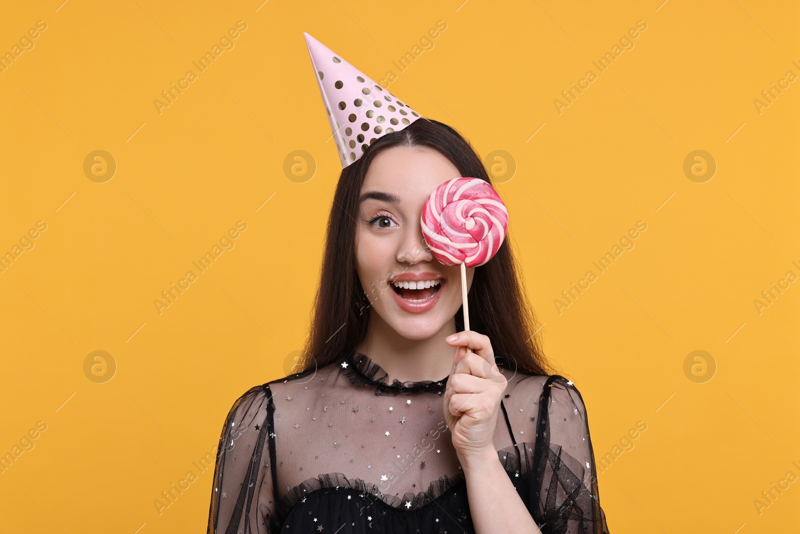Photo of Happy woman in party hat holding lollipop on orange background