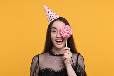 Photo of Happy woman in party hat holding lollipop on orange background