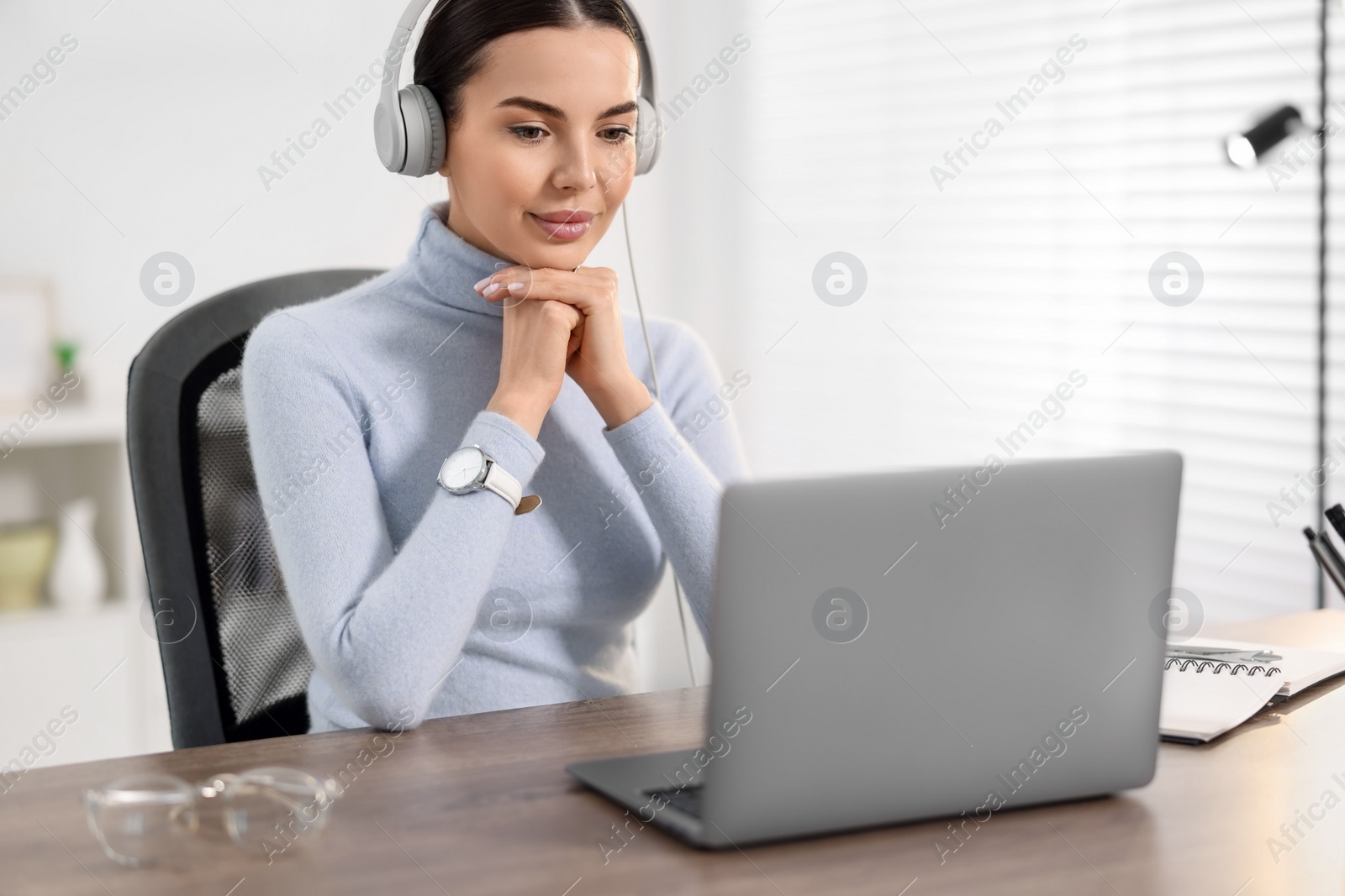 Photo of Young woman in headphones watching webinar at table in office