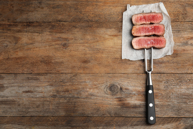 Photo of Carving fork with slices of delicious beef steak on wooden table, top view. Space for text