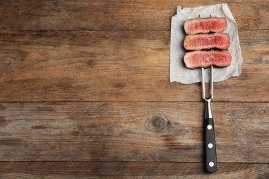 Carving fork with slices of delicious beef steak on wooden table, top view. Space for text