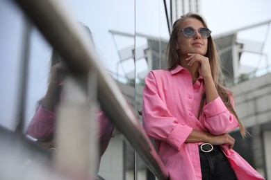 Beautiful young woman in stylish sunglasses near building outdoors, space for text