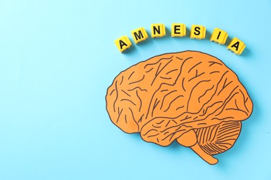 Photo of Yellow cubes with word Amnesia and paper cutout of human brain on light blue background, top view. Space for text