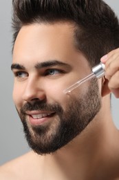 Handsome man applying cosmetic serum onto face on light grey background, closeup
