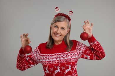 Photo of Happy senior woman in Christmas sweater and Santa headband holding festive baubles on grey background