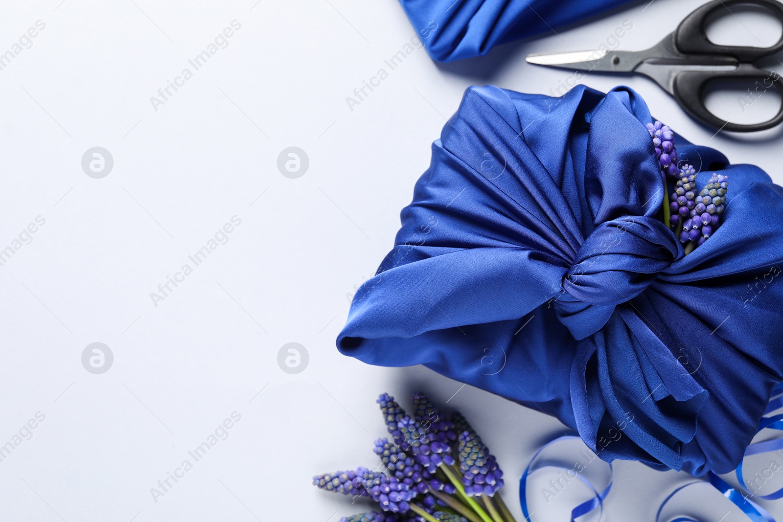 Photo of Furoshiki technique. Gifts packed in blue silk fabric, muscari flowers, ribbon and scissors on white table, flat lay. Space for text