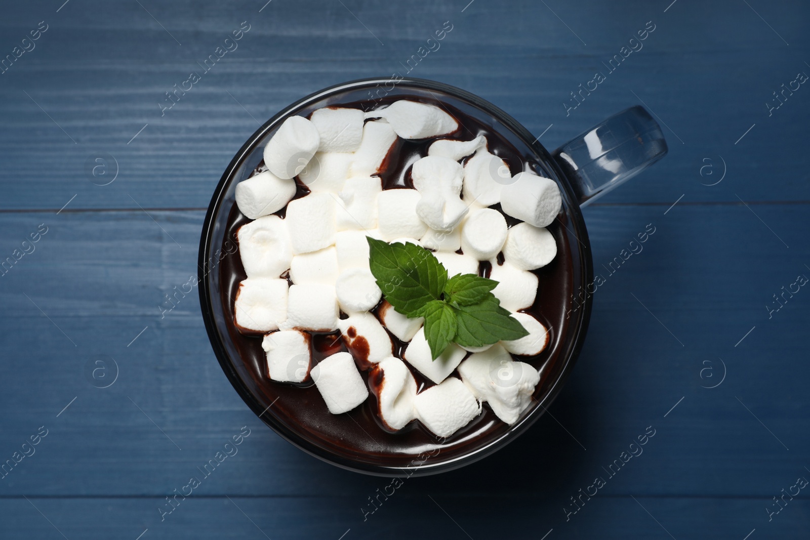 Photo of Glass cup of delicious hot chocolate with marshmallows and fresh mint on blue wooden table, top view