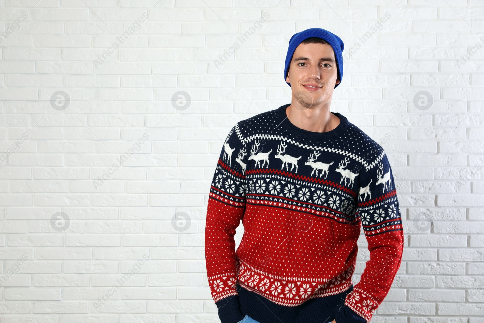 Photo of Portrait of young man in Christmas sweater and hat near white brick wall. Space for text